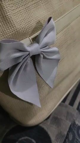 This video shows a close-up, static shot of a pale beige bag with a grey ribbon bow attached to it. The bag appears to be made of woven material with a textured surface. The bow is neatly tied and has two loops and two tails. The lighting is soft, casting a shadow on the lower left side of the bow. The background is out of focus, but appears to be a patterned surface, possibly fabric. There are no people or actions visible in the video. The video is very short, lasting only a few seconds.
