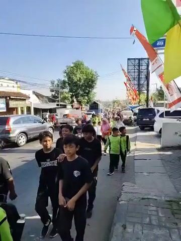 The video captures a procession moving down a street lined with buildings and trees. A group of young people, predominantly males, are walking in the foreground, some wearing black t-shirts and others green t-shirts. Several individuals are skateboarding. In the background, more people are walking, and a variety of vehicles, including cars, motorcycles, and a truck, are visible on the street. Colorful flags are strung across the street and attached to poles, adding a festive atmosphere. The sky is clear and blue, suggesting it is a sunny day. The scene appears to be a public event or parade.