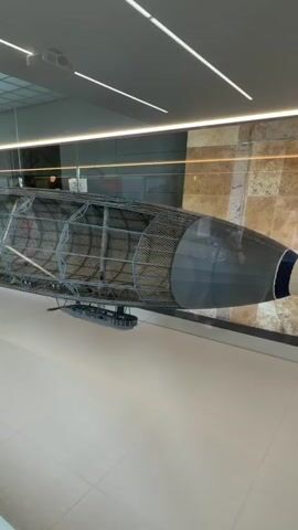 The video shows a large, partially constructed zeppelin-like object suspended indoors. The object has a metal skeletal frame with some fabric or material stretched across parts of it. The front section is covered in a dark gray material, with a blue and white band at the very tip. A small gondola-like structure is attached to the underside of the frame. The environment appears to be a museum or exhibition hall, with a tiled ceiling, spotlights, and stone-patterned walls in the background. The floor is a light color. The camera pans slowly along the length of the object.