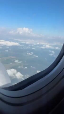 The video captures a static view from an airplane window, looking out over the landscape and sky. The upper portion of the frame shows a clear blue sky with scattered white cumulus clouds at varying altitudes. Below the clouds, a green and brown landmass is visible, suggesting a mix of vegetation and possibly some bare earth or agricultural fields. A portion of the airplane's engine or wing is visible in the lower left quadrant of the frame, appearing metallic and curved. The window frame itself occupies the right side and bottom of the video, showing its multi-layered structure. The scene is brightly lit, indicating daytime. No people or specific identifiable objects other than the aircraft parts and landscape are present.
