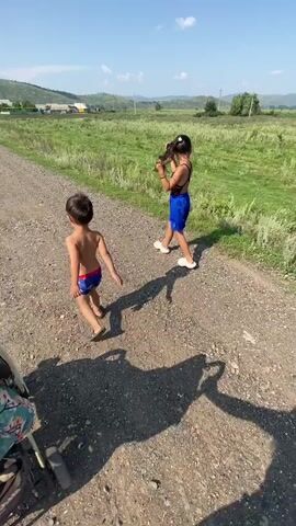 This video captures two children on a gravel path on a sunny day. In the foreground, a young boy, likely a toddler, is walking away from the camera. He is shirtless and wearing blue and red swim trunks. To his right and slightly ahead, a girl is walking in the same direction. She has long dark hair, which she is holding, and is wearing a black swimsuit top and blue shorts. Her hair is braided or tied back. They are both barefoot or wearing minimal footwear. A stroller is partially visible in the lower left corner of the frame, and a person's feet in purple sandals are in the lower right.

The background depicts a rural landscape under a clear blue sky with a few scattered clouds. Rolling green hills dotted with trees and some scattered buildings can be seen in the distance. The overall impression is of a warm, bright day in a country setting, possibly a village. The children appear to be enjoying an outdoor excursion.