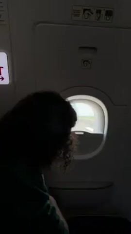 A person is silhouetted in an airplane seat, looking out the window. The view through the window shows an airport runway, with grass and sky visible in the distance. The image is taken from inside the airplane cabin.