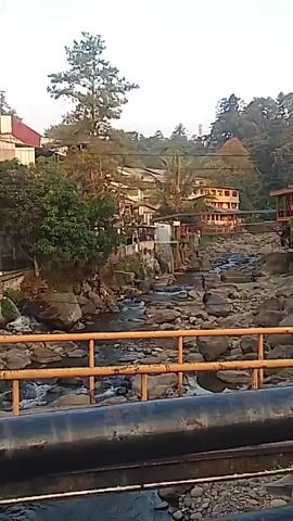 A video pans across a rocky riverbed with a shallow stream flowing through it. Buildings are visible on a hillside overlooking the river, with trees and foliage dotting the landscape. A yellow railing and two large black pipes are in the foreground, suggesting the perspective is from a bridge or elevated walkway. The sky is bright, indicating daytime. There are no people or animals visible in the video.