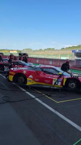 The video shows a pit lane at a race track. A red Ferrari race car is parked on the left, with its wheels removed and being worked on by pit crew members wearing red helmets. To the right of the Ferrari, several green Lamborghini race cars are lined up. These cars are connected to a fueling station, with hoses running from tanks to the vehicles. Pit crew members in yellow and black uniforms are attending to the Lamborghinis. In the background, grandstands filled with spectators are visible under a clear blue sky. The scene appears to be in preparation for or during a racing event.
