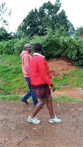 Two men are walking on a dirt road. The man on the left is wearing a pink hoodie and blue jeans. The man on the right is wearing a red jacket and plaid shorts. They are walking away from the camera. There are trees and bushes on either side of the road. The sky is overcast.