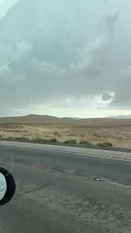 The video is a first-person perspective, likely from inside a vehicle, looking out at a desert landscape under a cloudy sky. The road is visible in the foreground, with tire marks and some wet patches suggesting recent rain. A side mirror of the vehicle is visible on the left. The middle ground shows sparse vegetation, dry grasses, and scrub brush. In the background, rolling hills or mountains are visible, partially obscured by the overcast sky. The overall impression is a vast, somewhat desolate environment with signs of impending or passing weather. There are no people or animals visible.