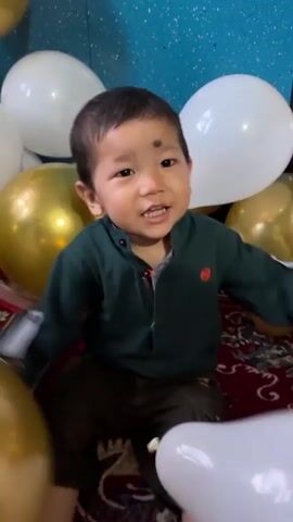 A child is sitting on a rug surrounded by white and gold balloons. The child is wearing a dark green jacket and green pants. The child is looking up and appears to be laughing or shouting.