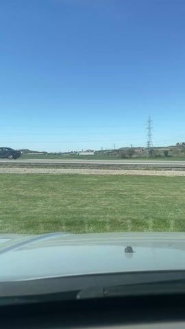 The video shows a view from inside a vehicle, likely a car, with the dashboard and windshield visible. Through the windshield, one can see a clear blue sky and a landscape featuring a stretch of green grass. Beyond the grass is a road with a guardrail, and cars can be seen driving on it. In the background, there are power lines and some indistinct buildings or structures. The video is stable, with the camera not moving significantly. There are no discernible people within the frame, only glimpses of passing vehicles.