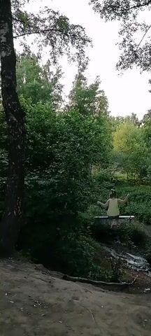 A young girl with blonde hair in a ponytail, wearing a light yellow dress and grey shorts, is swinging on a homemade swing. The swing appears to be made of wood and rope, suspended from a tree. The girl is actively swinging, her arms outstretched. The background is a lush, green forest with a dense canopy of trees. The ground in the foreground is sandy and covered with some debris. The lighting suggests it is daytime, possibly late afternoon, with a slightly overcast sky. The camera is positioned at a low angle, looking up at the girl and the swing.