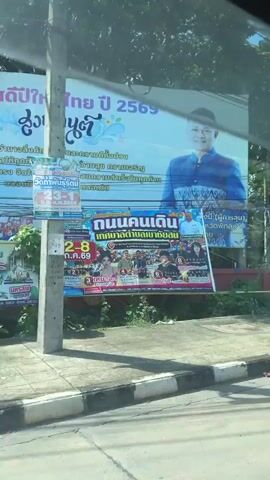 The video begins with a view of a large billboard featuring a portrait of a man in a blue uniform. Below this, another billboard displays images of people, likely politicians. The scene then transitions to a view from inside a vehicle, showing a wall decorated with Buddhist symbols and small stupas. The car continues on a road with other vehicles, passing under a pedestrian bridge. The sky is blue with scattered clouds, and trees and utility poles line the road.