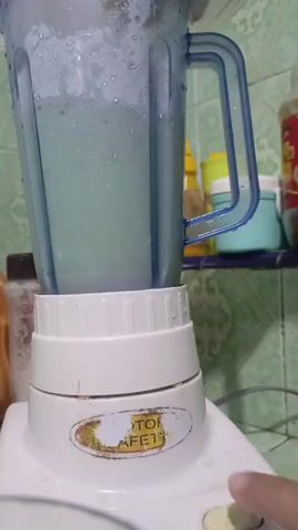 The video shows a close-up shot of a blender in use. The blender's pitcher is partially filled with a light green liquid. The pitcher is blue and clear plastic, and a blue handle is attached to its side. The base of the blender is white and has a circular sticker with the words "MOTOR SAFE" and a number "13" on it. A finger is pressing a button on the blender's base, indicating that it is operating. The background appears to be a kitchen with tiled walls and shelves with various containers and bottles. The lighting suggests it is indoors. The video captures the action of blending.