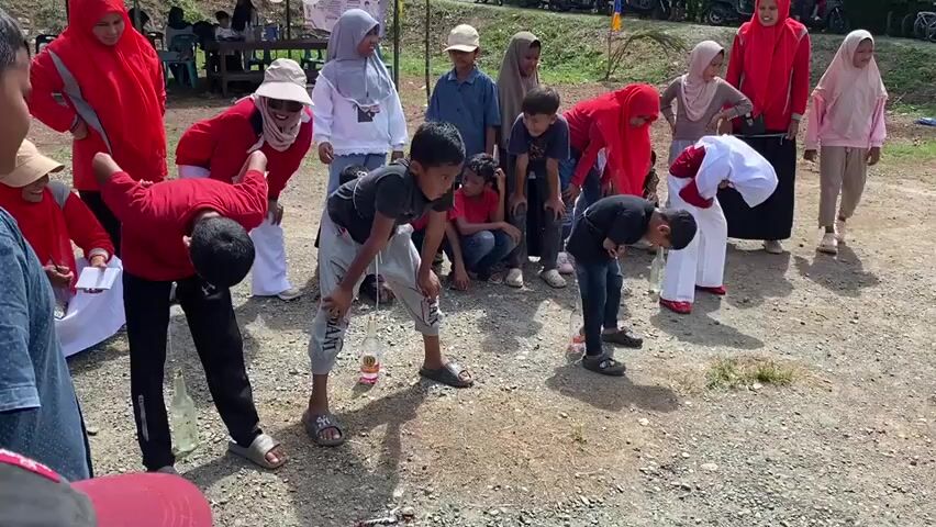 The video shows a group of children and adults gathered outdoors on a gravelly, dirt-like surface, possibly for a game or event. Several children are bent over, with bottles placed between their legs. Some adults, mostly women in red and beige hijabs, are watching the children. The scene appears to be daytime, with a bright sun. In the background, there are trees, some structures, and vehicles, suggesting an outdoor recreational area or a community gathering. Some of the children are holding or interacting with the bottles, possibly engaged in a challenge or competition. The overall atmosphere seems festive and engaging.