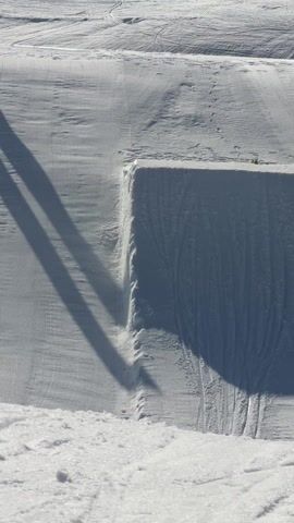 The video shows a skier performing a trick jump in a snowy, outdoor environment. The skier, dressed in black, approaches a snow-covered ramp, launches into the air, executes a backflip, and lands smoothly on the downward slope. The scene is set on a sunny day, and the crisp white snow contrasts sharply with the skier's dark attire. Another person stands off to the side, observing the jump, and a banner with "Alpe d'Huez" is visible in the background.