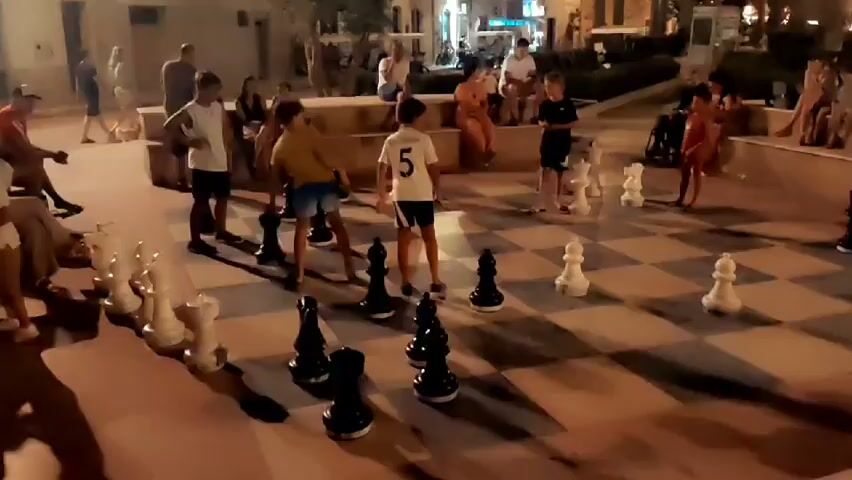 The video shows an outdoor scene at dusk or night, with people gathered around a giant chessboard. The chessboard is laid out on a paved surface with large, black and white chess pieces positioned on it. Several individuals, appearing to be mostly young people, are standing or walking around the chessboard, some appearing to be playing or observing a game of chess. Spectators are seated on tiered seating in the background. The environment appears to be a public square or plaza, possibly in a warm climate given the clothing of the people. The lighting suggests artificial illumination, possibly streetlights or ambient lighting from surrounding buildings, casting shadows on the ground. The overall atmosphere seems relaxed and social.