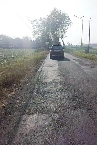 The video shows a dark-colored SUV driving on a paved road. The road appears to be in a rural or semi-rural setting, with trees and fields visible on either side. The sun is shining, casting long shadows. There are two utility poles on the right side of the road. The vehicle is moving away from the camera. The road surface is uneven in places, with visible patching and cracks. The overall impression is of a dry, sunny day.