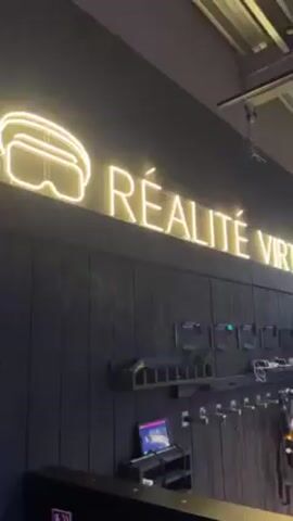 The video depicts the interior of a virtual reality gaming center. The entrance area features a neon sign that reads "Réalité Virtuelle," with rows of VR headsets and controllers stored below. The gaming area is visible through a glass partition, showing people wearing VR headsets and holding gun-shaped controllers, presumably engaging in a virtual shooting game. The room is lit with neon lights in various colors, and geometric symbols are placed on the floor and walls. People are moving around, suggesting active participation in the virtual reality experience.