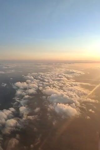 The video shows an aerial view from an airplane window. The camera is pointed downwards and towards the horizon, capturing a vast expanse of clouds. The sun is setting or rising, casting a warm, golden light on the clouds and the sky. The lower part of the frame shows a portion of the airplane's engine. The scene is peaceful and serene, with the clouds forming a textured landscape beneath the aircraft.