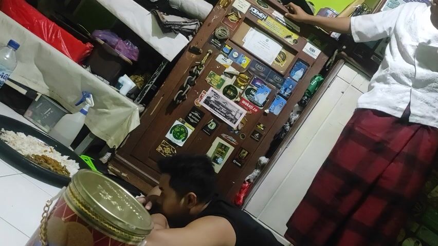 The image shows a low-angle shot of a small room. A man in a black tank top lies on the floor facing a wooden cabinet covered in stickers. To his left is a plate of food and a container with a gold lid and chain. Another person, wearing a white shirt and red plaid sarong, stands near the cabinet. The room is cluttered with items on shelves and the floor, including a water bottle and cleaning supplies. The lighting is dim and the composition is slightly off-kilter.