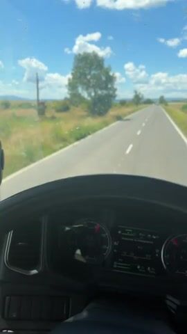 The video provides a first-person perspective from inside a truck cab, looking out through the windshield and a side mirror. The truck is driving on a paved road through a rural landscape under a bright, cloudy sky. The road stretches ahead, with a grassy field and scattered trees on either side. The dashboard of the truck is visible in the foreground, showing gauges and a digital display. The side mirror reflects the road and scenery behind the truck, including a solitary tree and power lines. The overall impression is of a daytime journey through an open, sunlit countryside.