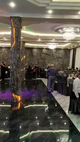 The video appears to show an indoor event, possibly a wedding reception or a formal dinner. The setting is a banquet hall with elegant decor, including chandeliers, ornate ceiling designs, and stone-like wall coverings. The room is filled with round tables draped in grey tablecloths, set for dining. Several people are visible, some seated at the tables and others standing and mingling. In the foreground, a large decorative pillar with a rough, stone-like texture and illuminated with what appears to be colored lighting is prominently featured. The floor is highly polished, reflecting the lights and surrounding decor. Toward the back of the room, there is a stage area with a cake displayed on a table, adorned with roses. A man in a white shirt is standing near the stage, and other individuals are seated behind him. The lighting in the room is a mix of ambient and accent lighting, with colorful projections visible on a white curtain in the background. The overall atmosphere seems to be one of a celebratory gathering.