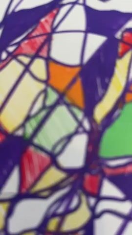 The video shows a close-up view of a piece of abstract art, rendered on white paper with various colored markers. The drawing consists of a complex network of interwoven lines and shapes, filled with vibrant colors including purple, orange, green, yellow, and red. Some sections are filled solid, while others are hatched with parallel lines. The camera then slowly pulls back, revealing that the artwork is on a wooden table. Several marker pens in different colors are scattered around the artwork. In the background, a glass bottle of San Pellegrino water, a glass of water, a stack of blue napkins in a glass container, a smartphone, and some papers are visible on the table, suggesting a casual setting, possibly a cafe or a home. The video is shot from a slightly elevated angle, with the focus initially on the artwork and gradually expanding to the surrounding environment.