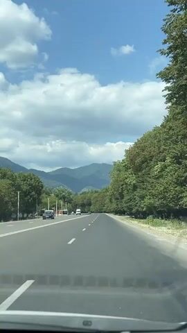 The video shows a view from inside a vehicle driving on a paved road. The road has lane markings and some cars are visible. On the left side of the road, there are some trees and a white structure that appears to be a toll booth or checkpoint. On the right side of the road, there is a dense line of trees. In the distance, there are mountains under a bright blue sky with scattered clouds. The camera is facing forward, capturing the road ahead and the surrounding landscape. The video is short, showing the car moving along the road for a few seconds.