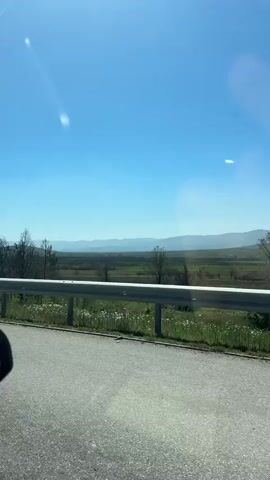 The video shows a view from a moving vehicle, likely a car, driving along a paved road. A gray metal guardrail separates the road from a grassy area with wildflowers. Beyond the grass and scattered, mostly leafless trees, there is a large, open field. In the far distance, hazy mountains are visible against a clear, bright blue sky. Sunlight is reflecting off the car's windshield.
