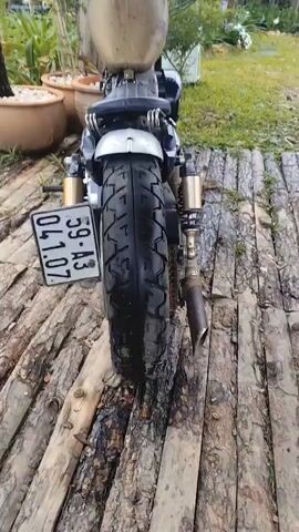 The video shows a close-up view of a custom motorcycle from the rear, moving forward to reveal the front. The motorcycle has a black tire with a unique tread pattern, a silver fender, and gold-colored shock absorbers. A license plate with Vietnamese characters is visible. The motorcycle is parked on a wooden platform outdoors, with potted plants and greenery in the background. The fuel tank is a tarnished gold color, and the seat is black and appears to be made of leather. The handlebars are chrome and feature black grips and controls. The video is a stationary shot that pans slightly to show different angles of the motorcycle.