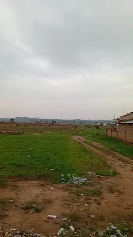 A video shows a partially developed rural area under a cloudy sky. A dirt road with scattered trash runs through the foreground. To the right, a brick wall with a building behind it marks a property line. The middle ground is a grassy field with construction materials, mounds of dirt, and scattered buildings. In the background, hills and more buildings are visible under the overcast sky.
