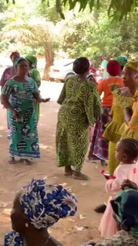 The video shows a group of women dancing in an outdoor setting. They are wearing colorful, patterned dresses and headscarves. Some are standing and watching, while others are dancing. A child in pink is also present. The setting appears to be a sandy area with trees providing shade, possibly a rural or village environment. A car is visible in the background. The focus is on the dancing and the festive atmosphere.
