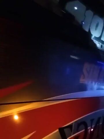 The video shows the rear and side of a red bus at night. The bus has a distinctive design with black and red accents. On the rear of the bus, text reads "SINAR MUDA" and "SCANIA K410". Lights are visible on the bus and in the background, indicating it is on a road. The bus appears to be in motion.