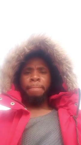 A close-up shot shows a man with a dark beard, wearing a gray shirt and a bright red coat with a fur-lined hood. He is speaking and looking directly at the camera. The background is a blurred white sky with a hint of vegetation at the bottom.