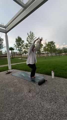 A woman in athletic wear is performing a yoga pose on a mat outdoors. She is in a chair pose with her arms raised and then twists her torso to the side, bringing her hands together in front of her chest. She is positioned under a modern, outdoor structure with a metal roof and frame. The background shows a green lawn and trees under a cloudy sky. A water bottle and a portable speaker are on the ground next to the mat.