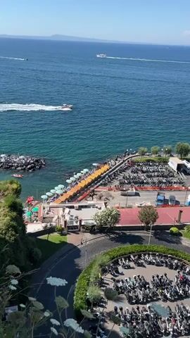 The video shows a panoramic view of a coastal area with a body of water, likely the sea, on the right side of the frame. In the foreground, there is a curved road with a circular parking area filled with numerous motorcycles. Beyond the road, there is a strip of land adjacent to the water, featuring a row of lounge chairs and umbrellas set up on what appears to be a beach or a seaside resort. This area also contains a parking lot with cars and several buses. In the distance, the sea stretches out, with a few boats visible, and a mountainous landmass or volcano is present on the horizon. The sky is clear and blue, indicating a sunny day. The overall scene depicts a bustling tourist destination with a focus on leisure and transportation infrastructure.