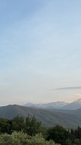 The video shows a landscape featuring rolling green hills in the foreground and a mountain range in the background. The mountain peaks are snow-capped and partially obscured by a layer of clouds. The sky is a pale blue, with some thin cloud cover present. Some trees are visible in the foreground, partially obscuring the lower part of the hills. The lighting indicates it is either early morning or late afternoon.