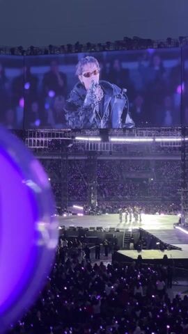 Here is a description of the video:

The video shows a performer on a large screen above a stage in a stadium. The stadium is filled with people holding lights, mostly purple. The performer on the screen is wearing sunglasses and a black outfit with silver chains. He is holding a microphone and appears to be singing. On the stage below, other performers are visible. The stage is lit up with bright lights. The overall atmosphere is energetic and vibrant.