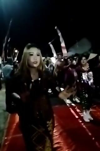 A nighttime outdoor scene shows a group of people dancing. The foreground features a woman in a dark, patterned outfit, possibly traditional attire, with blonde hair and light makeup. She is extending her right hand, palm facing forward, as if to stop something or someone. Behind her and to her right, other individuals are also dancing or in motion. They are dressed in various colorful garments, some appearing to be traditional clothing. Red fabric is laid out on the ground in front of the dancers, possibly serving as a stage or walkway. Flags, some with red and white stripes, are visible in the background, suggesting a celebratory event or cultural gathering. The lighting is dim, with some artificial light illuminating the area.