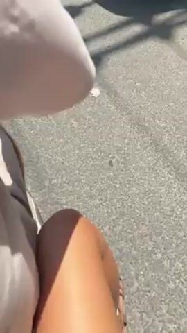 The video shows a person riding a motorcycle down a paved road. The camera is mounted on the motorcycle, providing a first-person perspective. The person is wearing a white shirt and shorts, and their leg is visible in the lower portion of the frame. The road has parked cars, trees, and buildings on either side, typical of a residential or commercial area. The weather appears to be sunny. A side mirror of the motorcycle is also visible in the frame. The road appears to be generally clear of heavy traffic.