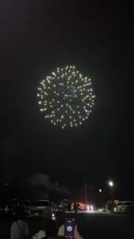 At night, people stand in a parking lot watching fireworks explode in the sky above. The fireworks are colorful, with bursts of white, yellow, and orange. Some people are holding up their phones to record the fireworks. The environment includes parked cars, street lights, and trees. The atmosphere is celebratory.