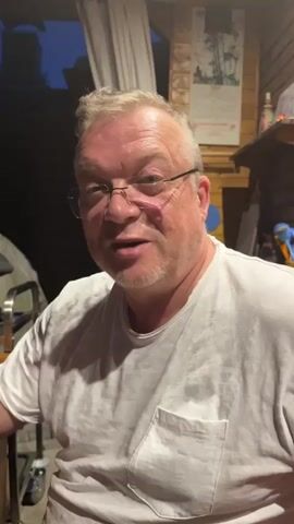 A man with glasses and a white t-shirt is looking directly at the camera and speaking. He has short, light-colored hair and a goatee. The background appears to be outdoors, possibly a patio or porch, with wooden walls and a white curtain visible. There are also some objects on a shelf in the background, including what looks like a picture and some bottles. The lighting suggests it is either evening or early morning. The man is smiling slightly.