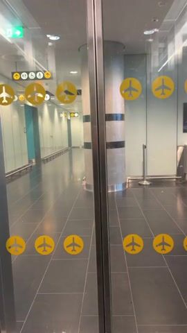 The video shows a view through a set of glass doors. The doors appear to be in an airport or a similar large public building. There are yellow circular stickers with airplane silhouettes on the glass doors. On the wall in the background, there are signs with directional arrows and symbols indicating amenities like restrooms and a flight information display. The hallway beyond the doors is lined with pillars and appears to be an access route.
