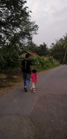 Two people, an adult and a child, are standing on a paved road in a rural area. The adult, wearing a black t-shirt and blue jeans, is on the left, and the child, wearing a pink shirt and light-colored pants, is on the right. They appear to be interacting, possibly holding hands. The road is surrounded by vegetation, including trees and bushes, and a small building is visible in the background. The sky is overcast. The video is filmed from a slightly elevated perspective and is rotated 90 degrees counterclockwise.