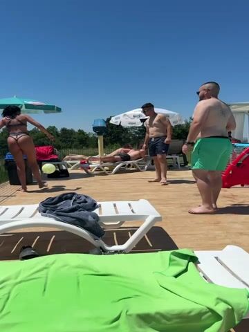 A group of people are playing with a ball on a wooden deck under a clear blue sky. A woman in a bikini kicks the ball towards a man in green shorts. Another man stands between them, watching. A third man lies on a lounge chair in the background. There are lounge chairs and umbrellas scattered around the deck. The overall atmosphere appears to be a relaxed, outdoor gathering.