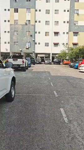 A white truck is parked in a lot in front of a multi-story apartment building. The building has white, yellow, and grey sections. There are several other cars parked in the lot, including a blue sedan, a black SUV, and a white car. The lot is paved and has white dashed lines marking parking spaces. Trees are visible behind the building on the right side of the frame.