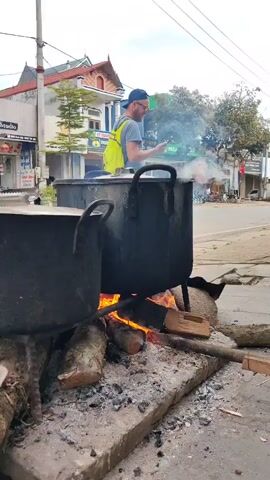 Here is a description of the video:
The video shows a roadside cooking setup in a Vietnamese town.  Two large, black metal pots sit atop a makeshift hearth of concrete slabs and burning wood. Flames are visible beneath the pots. A man in a grey t-shirt, khaki pants, and sunglasses walks by, glancing at his phone and then taking a photo of the setup. The background includes buildings with Vietnamese signage, power lines, a Vietnamese flag, and light vehicle traffic on the street. The sky is overcast.