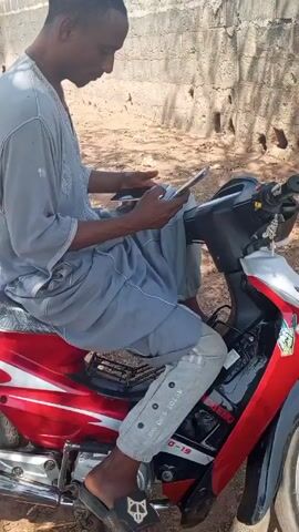 A young Black man sitting on a red and black motorcycle, looking at his smartphone, outdoors during the day. The man is wearing a long gray shirt and gray pants. He is sitting on the motorcycle, which is parked on what appears to be a dirt driveway next to a concrete block wall. Music is playing in the background.