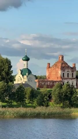The video shows two churches situated on a grassy hill overlooking a body of water. The church on the left is white with a green dome and a cross on top. The church on the right is in ruins, made of red brick with a crumbling dome. Trees and reeds surround the churches, and the sky is blue with some clouds. The water is calm and reflects the sky. The lighting suggests it is either early morning or late afternoon, with the sun casting a warm glow on the scene.