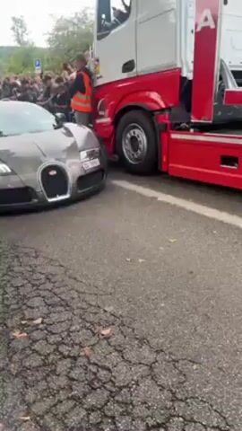 A tan Bugatti Veyron with a rear spoiler and Swiss license plate TG-78551 is parked on a cracked asphalt road next to a red and white flatbed truck. A crowd of spectators surrounds the scene, some taking photos. Buildings with windows are visible in the background. The car's passenger door is open, and a person is sitting in the driver's seat.