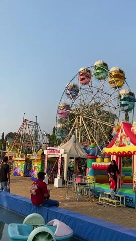 The video shows an outdoor amusement park with various rides. In the background, a large Ferris wheel with colorful gondolas is visible, as well as a smaller roller coaster-like ride. In the foreground, there is a large inflatable playground with slides and climbing structures, adorned with cartoon characters. A woman in a black headscarf and a young child in a red shirt are standing on a platform of the inflatable playground. To the left, a man wearing a red t-shirt with a graphic design is sitting on the edge of a blue barrier, facing away from the camera. Other people are scattered around the area, and trees are visible in the distance. The ground appears to be dirt and gravel. The sky is clear and blue.