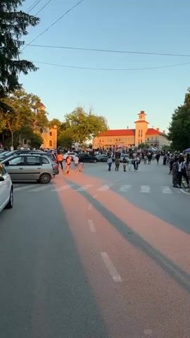 The video pans across a street scene with a crowd of people gathered. The sun is setting, casting long pink shadows across the road and illuminating the buildings in the background. Cars are parked on the left side of the street. In the center of the road, a crosswalk is visible. People are walking across the street and gathering in groups. There are trees and buildings in the background. The sky is clear and blue.
